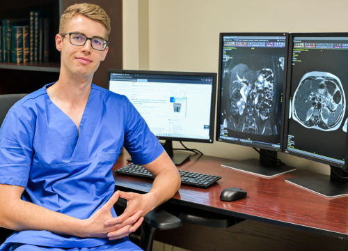 Młody mężczyzna w okularach, ubrany w niebieski fartuch lekarski. Siedzi przy stole. Na stole są trzy ekrany komputerowe. Na dwóch wyświetlają się obrazy radiologczne.