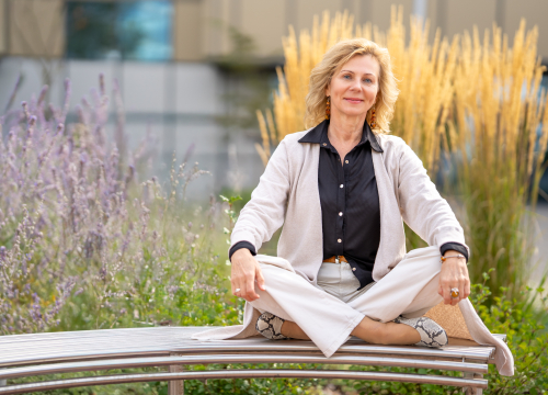 Kobieta siedzi na ławce w ogrodzie, mając skrzyżowane nogi. Ubrana jest w jasne spodnie, czarną koszulę i jasny sweter. W tle widoczne są rośliny ozdobne.