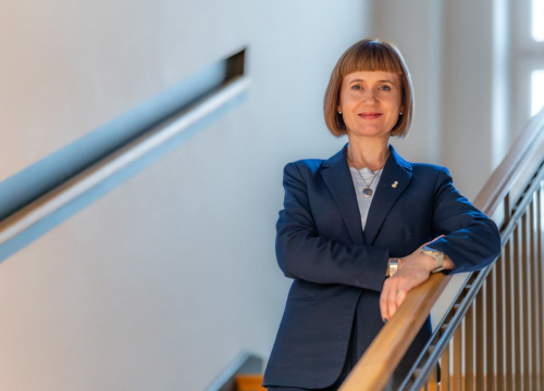 Kobieta w granatowej marynarce stoi na schodach oparta o poręcz.