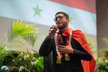 Młody chłopak w okularach z flagą Syrii na ramionach śpiewa do mikrofonu.