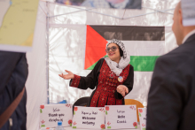 Młoda dziewczyna z "arafatce" na głowie i tradycyjnym stroju. Wykonuje gest zapraszający do stoiska. Za nią flaga Palestyny.