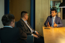 Three men. They are sitting at a table in an office room. They are all dressed in suits. One of them, on the right, facing the camera. He has glasses and a beard. He is saying something to the others. The two men look at him.