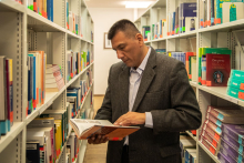 A man in a library room, between bookshelves. He stands and browses intently through a book.