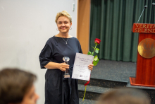 A woman smiling for a photograph. In her hand she holds a rose, a diploma and a statuette.