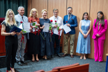 Group of people. They are standing in the hall. They are smiling at the camera. Some of them are holding flowers and diplomas.