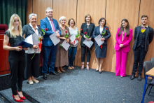 Group of people. They are standing in the hall. They are smiling at the camera. Some of them are holding flowers and diplomas.