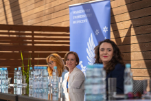Trzy kobiety siedzące przy stole. Przed nimi, na stole stoją butelki z wodą i szklanki. Za nimi, po lewej stronie stoi rollup granatowo-niebieski z orłem WUM.