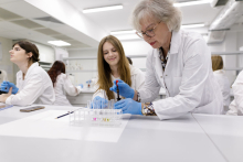 Dwie młode dziewczyny i kobieta w starszym wieku w białych fartuchach w laboratorium analitycznym.