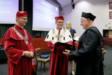 Dwóch mężczyzn w czerwonych togach wręcza dyplom mężczyźnie w czarnej todze.