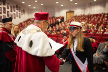 Mężczyzna w średnim wieku ubrany w togę rektorską ściska dłoń młodej dziewczynie, ubranej elegancko, z czerwono-białą szarfą i czapką akademicką.