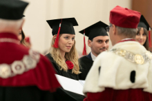 Grupa osób ubranych w czarne togi akademickie oraz mężczyzna ubrany w togę białą-czerwoną oraz mężczyzna ubrany w biało-czarną togę, wręczają dyplomy.