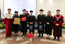 Grupa młodych osób ubranych w czarne togi akademickie stoi z dyplomami w rękach, uśmiecha się do aparatu. Obok rektor ubrany w czerwono-białą togę akademicką oraz dziekan ubrany w togę czarno-czerwoną.