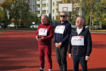 Three men dressed in sportswear. On their sweatshirts they have cards with the names of those who fought for Ukraine stuck to them. The men are standing on the school field.