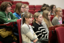 Grupa młodych osób siedzi na czerwonych fotelach i patrzy przed siebie