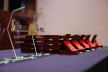 Trzy przezroczyste szklane trofea z napisami oraz kilka czerwonych pudełek zawierających złote medale ułożone w rzędzie. 