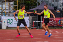 Two runners in yellow shirts are participating in a relay in the stadium. The athlete with the number 1060 passes the baton to the runner with the number 1067. One is wearing pink, the other blue shoes. In the background are grandstands, tents and banners.