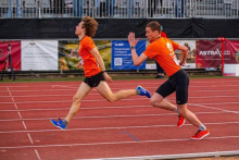 Two men in orange T-shirts and black shorts are running on a red athletics treadmill. One of them is slightly in front. In the background you can see grandstands with barriers and advertising banners.