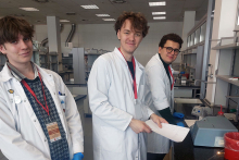 Trzech młodych mężczyzn stojących w laboratorium. Wszyscy mają na sobie białe fartuchy laboratoryjne i czerwone identyfikatory zawieszone na szyi. Jeden z nich trzyma w ręku kartkę papieru. W tle znajdują się różne urządzenia laboratoryjne oraz meble, takie jak stoły i szafki.