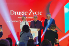 Trzy osoby stojące na scenie. Dwie z nich trzymają plakat. W tle znajduje się czerwone tło z białym napisem: „Drugie Życie” oraz „Aktywni dla transplantacji”. Na pierwszym planie widać fotografa robiącego zdjęcia.