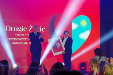 Dwie osoby stojące na scenie podczas ceremonii wręczenia nagród. Jedna osoba trzyma oprawiony obraz, druga klaszcze. W tle znajduje się duży czerwony ekran z białym napisem: „Drugie Życie – Nagroda w kategorii Osobowość polskiej transplantologii”. Na ekranie widoczna jest także grafika w kształcie serca w kolorach niebieskim i żółtym.