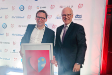 Dwie osoby w formalnych strojach stoją na tle ścianki z logotypami. Uśmiechają się do zdjęcia. Jedna osoba trzyma oprawiony obraz-dyplom. Na obrazie widnieje serce z grafiką przedstawiającą błękitne niebo i żółte słońce. 