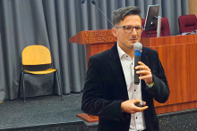 Młody mężczyzna stojący na scenie sali konferencyjnej. W prawej ręce trzyma mikrofon. Ma na sobie czarną marynarkę i białą koszulę. W lewej ręce trzyma pilot do prezentacji. Za nim stoi drewniana mównica. W tle widać szare zasłony, żółte krzesło i pulpit z komputerem.