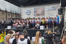 Na scenie są osoby w różnym wieku. Niektóre osoby trzymają mikrofony. Inne mają na sobie koszulki z numerami startowymi. Przed sceną stoi grupa fotografów i kamerzystów, którzy robią zdjęcia i nagrywają wydarzenie. W tle znajduje się baner z napisem: „Bieg po Nowe Życie”. 