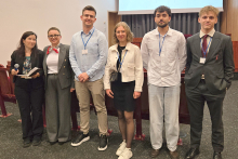 Sześć osób stoi w rzędzie w sali konferencyjnej. Są to studenci i organizatorzy konkursu. Część z nich jest ubrana formalnie, część nieformalnie. W tle widać ekran projekcyjny i ciemnoszare zasłony.