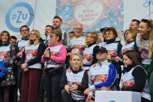 Grupa osób pozujących razem do zdjęcia. Większość z nich ma na sobie stroje sportowe i koszulki z kolorowymi wzorami i napisem: „Bieg po nowe życie”. W tle widoczny jest duży baner z tym samym napisem umieszczonym w kształcie serca. 