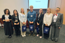 Siedem osób, sześć kobiet i mężczyzna, stoją w rzędzie w sali konferencyjnej. Osoby w środku, studenci, trzymają niebieskie torby z białym logo. Część osób jest ubrana formalnie, część nieformalnie. W tle widać ekran projekcyjny. Na ekranie znajduje się napis „Thank you” na białym tle.