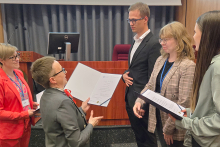 Pięć osób, cztery kobiety i mężczyzna, stoją w sali konferencyjnej zwróceni do siebie twarzami. Wszystkie osoby są ubrane formalnie. Po lewej stronie stoi osoba w czerwonym garniturze. Obok niej stoi osoba w szarym garniturze. Są to organizatorki konkursu, które wręczają dyplomy studentom. Pozostała trójka to studenci, uczestnicy konkursu. Są formalnie ubrane. W tle widać drewnianą mównicę i ekran projekcyjny zasłonięty szarymi zasłonami.