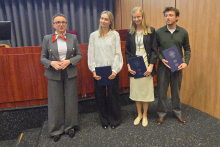 Cztery osoby stojące w rzędzie. Trzy osoby trzymają niebieskie teczki. Pierwsza osoba z lewej ma szary garnitur i czerwoną apaszkę. Druga osoba nosi białą bluzkę i czarne spodnie. Trzecia osoba ma jasną spódnicę, ciemną marynarkę i żółty identyfikator na szyi. Czwarta osoba ma zielony sweter i szare spodnie. Tło stanowi drewniana ściana i zasłony. 