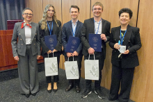 Pięć osób, trzy kobiety i dwóch mężczyzn, stoją w rzędzie i uśmiecha się do zdjęcia. Tło stanowi drewniana ściana. Każda osoba trzyma dyplom i białą torbę. Wszystkie osoby są ubrane elegancko, w garnitury i marynarki. 