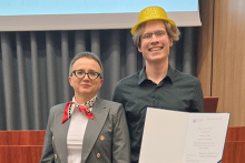 Kobieta i mężczyzna stojący koło siebie. Kobieta stoi po lewej i ma szary garnitur, białą koszulę i czerwoną apaszkę. Mężczyzna po prawej ma czarną koszulę i złoty kapelusz. Trzyma dokument w rękach. Dokument jest częściowo widoczny. W tle widać ciemnoszarą zasłonę i fragment ekranu projekcyjnego.