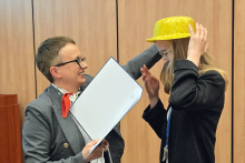 Po lewej kobieta, ubrana w szarą marynarkę z czerwoną apaszką na szyi, w prawej ręce trzyma otwartą teczkę. Zakłada na głowę kobiety po prawej złoty kapelusz. Kobieta po prawej ma jasne włosy i czarną marynarkę. Tło stanowi drewniana ściana z pionowymi panelami. 