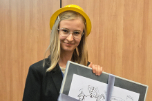 Młoda kobieta uśmiechająca się do zdjęcia. Ma na sobie czarną marynarkę i żółty kapelusz. Ma jasne włosy i okulary. Trzyma w rękach oprawiony rysunek z postaciami i tekstem. Rysunek jest przewiązany szarą wstążką. Tło stanowi drewniana ściana z pionowymi panelami. 