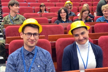 Mężczyzna i kobieta siedzący w czerwonych fotelach w sali konferencyjnej. Mają na głowie żółte czapki, a na szyi niebieskie smycze z identyfikatorami. W tle widać rzędy pustych czerwonych foteli oraz kilka innych osób, które na nich siedzą.