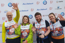 Cztery osoby stojące przed ścianką z logotypami i tekstem. Mają na sobie koszulki z numerem 23 i napisem „BIEG PO NOWE ŻYCIE”. Koszulki mają kolorowe wzory. Wszyscy mają na szyjach medale. Niektóre osoby machają rękami.