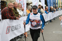 Prof. Danielewicz - uczestnik biegu z kijkami do nordic walking. Osoba ma na sobie koszulkę z numerem startowym. W tle stoją kibice za barierkami. Widoczne są także banery reklamowe.