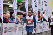 Prof. Mariusz Kuśmierczyk, uczestnik marszu z kijkami do nordic walking. Ma na sobie koszulkę startową z numerem. W tle znajdują się banery i flagi z różnymi logotypami i napisami. Za barierkami stoją kibice, którzy obserwują wydarzenie.