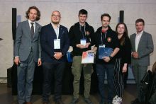 Pięciu mężczyzn i kobieta stoi w rzędzie na tle jasnej ściany. Trzy osoby po lewej mają garnitury, trzy po prawej czarne bluzy z logo. Osoba w środku trzyma trofeum, a osoba obok niej dyplom. Wszyscy mają identyfikatory na szyjach. Uśmiechają się do zdjęcia.