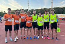 Seven men wearing athletic shirts with starting numbers stand on the treadmill of the athletics stadium. They are wearing orange or neon yellow T-shirts and black shorts. They stand close together, embracing each other with their arms. In the background are grandstands and trees.