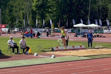 A female athlete in a yellow T-shirt and black shorts performs a long jump in the stadium. She is hovering over a sand pit with one hand up. Visible in the background are the referees, the competitors, the greenery and the stands with flags and organizers' tents.