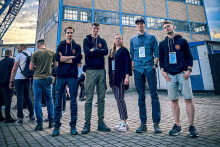 Czterech mężczyzn: dwóch po lewej i dwóch po prawej i kobieta w środku stoją na zewnątrz przed budynkiem z cegły i szkła. Mają identyfikatory na smyczach, twarze są zamazane. Trzy osoby w ciemnych bluzach, jedna w niebieskiej koszuli, jedna w czarnej bluzce i pasiastą spódnicą. W tle widoczni inni ludzie i metalowa konstrukcja.