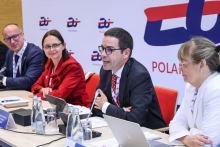 Four people are seated at a conference table. The men are wearing navy blue jackets and white shirts. The man sitting second from the right speaks into a microphone. The first from the right sits a woman wearing a white jacket. She looks ahead. From the left sit a man and a woman. The woman is wearing a red jacket. Both are looking at the speaker. On the table are microphones, water bottles, cups and nameplates.