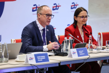 A woman and a man are seated at the table.  The man on the left is wearing a dark suit and white shirt, while the woman on the right is wearing a red outfit. On the table are microphones, water bottles, cups and nameplates.
