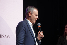 Mężczyzna w średnim wieku. Nosi granatową marynarkę i białą koszulę. Trzyma mikrofon przy ustach. Tło jest ciemne.