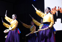 Grupa młodych kobiet, które tańczą na scenie. Kobiety mają na sobie żółte koszule i fioletowe spódnice. Każda z nich trzyma bębenek i pałeczki. Jedna ręka jest uniesiona z pałeczką, druga trzyma bębenek. Osoby stoją w rzędzie. Tło jest ciemne. Po prawej stronie widać fragment ekranu.