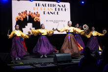 Pięć tancerek wykonujących tradycyjny koreański taniec bębnowy. Noszą żółte bluzki i fioletowe spódnice. Każdy trzyma bębenek i pałeczki. W tle widać duży ekran. Na ekranie jest napis: „TRADITIONAL KOREAN DRUM DANCE”. Na scenie stoją głośniki i sprzęt techniczny. Przed sceną siedzą widzowie. Jeden z widzów robi zdjęcie lub nagrywa telefonem.