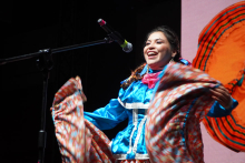 Kobieta stojąca na scenie za mikrofonem. Ma ciemne włosy. Uśmiecha się i śpiewa. Nosi niebieski strój z białymi ozdobami. Ma różową chustkę na szyi. Trzyma kolorową spódnicę i rozkłada ją na boki. Tło jest ciemne. Po prawej stronie widać duży pomarańczowy wzór. 
